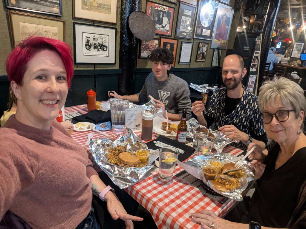 A group of four people enjoying a meal at a barbecue restaurant, seated at a table with checkered tablecloths, surrounded by framed pictures and memorabilia on the walls.