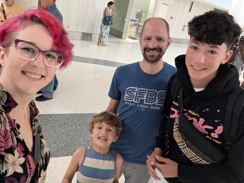 A group selfie taken at an airport, featuring a woman with pink hair and glasses, a man with a beard, a smiling young boy, and a teenage boy wearing a black hoodie with colorful text.