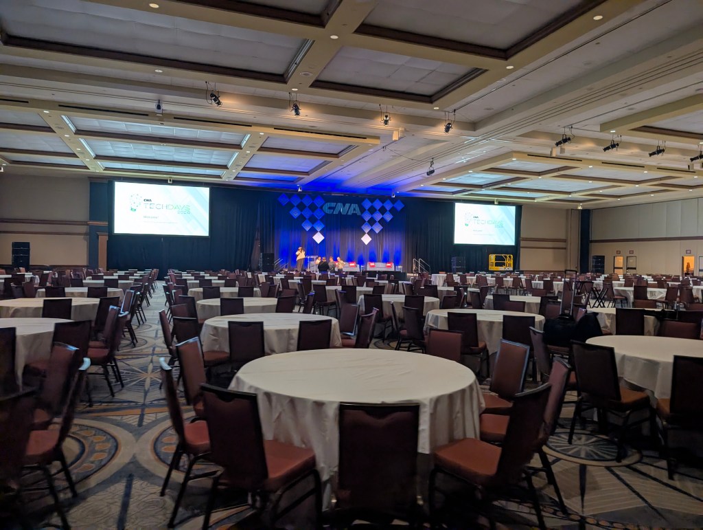 A spacious conference hall set up for an event, featuring multiple round tables with white tablecloths, empty chairs, and a stage adorned with blue lighting and presentation screens displaying 'CMA Tech Days 2023'.