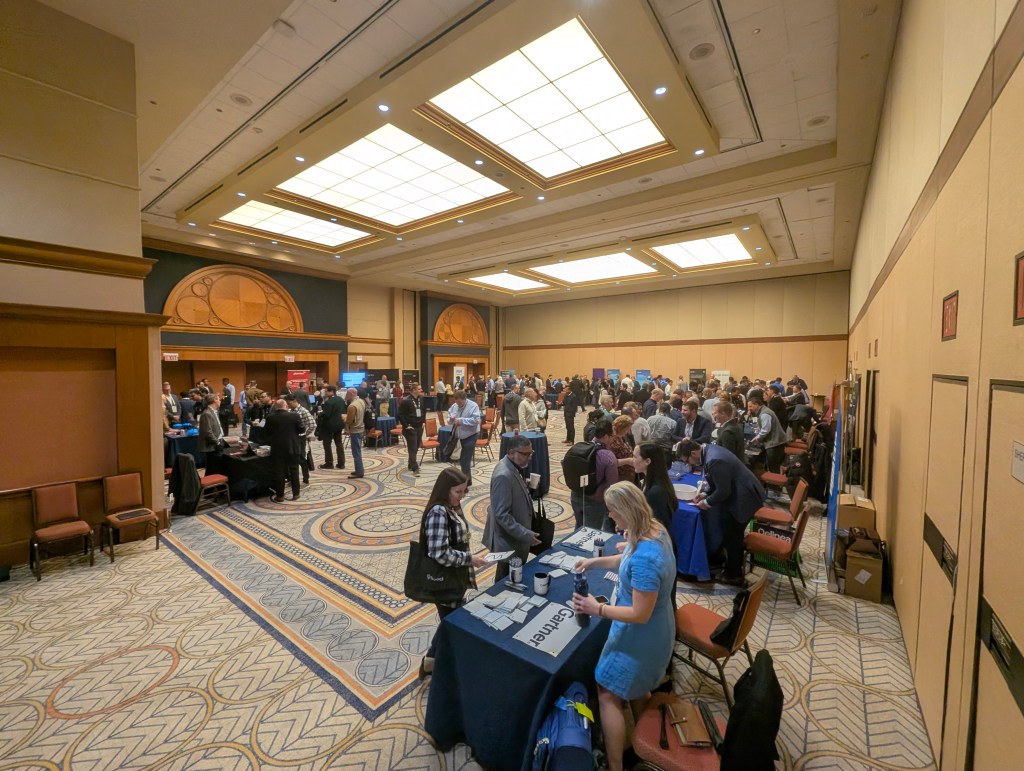 An indoor convention or conference setting with many attendees networking at various booths. Tables are set up with displays, and there are groups of people engaged in conversation throughout the room.
