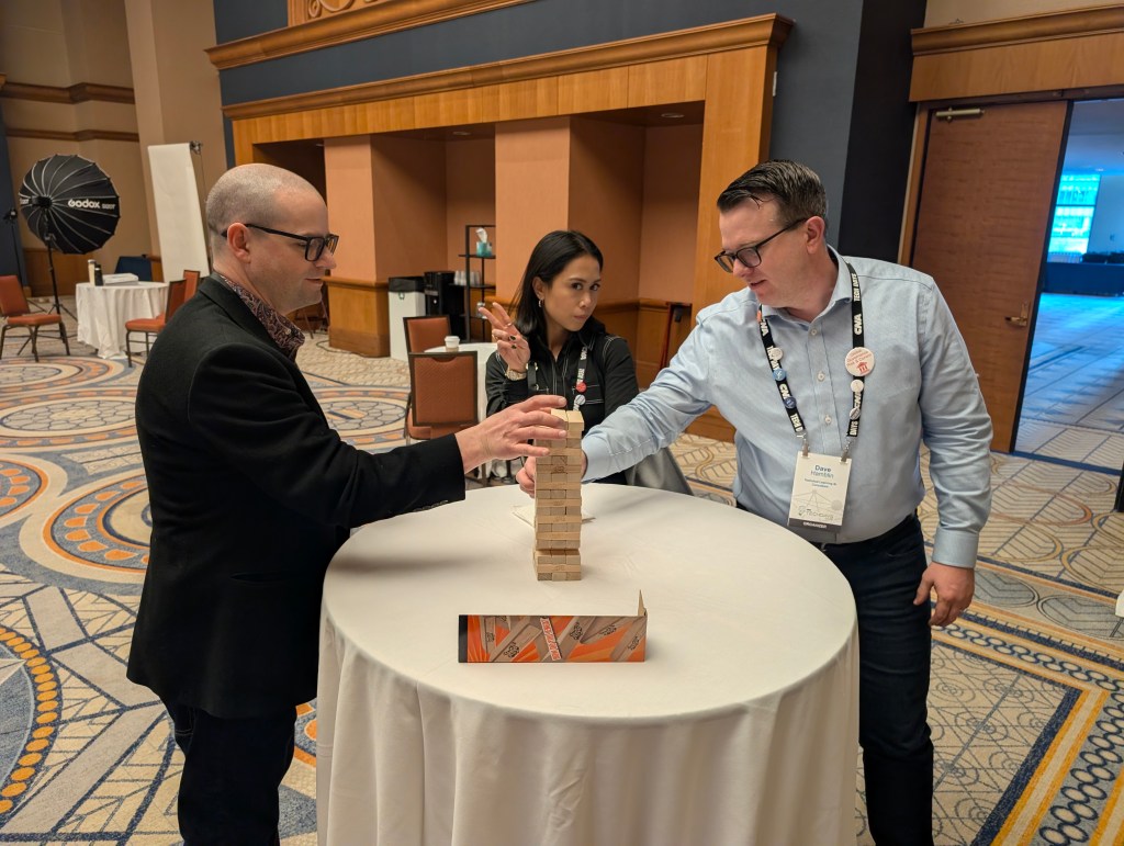Three people interacting around a table with a Jenga-like game, one person pulling a block, another observing, and the third person preparing to take their turn.