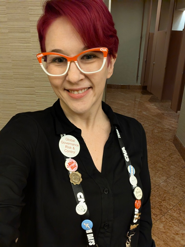 A smiling person with pink hair wearing orange-framed glasses and a black shirt, showcasing a lanyard with various badges and buttons, including one labeled 'Global Governance Risk & Control.'