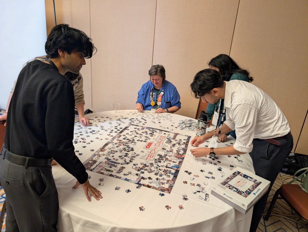 A group of people gathered around a table working on a jigsaw puzzle during an event, with pieces spread out and some individuals focused on fitting pieces together.