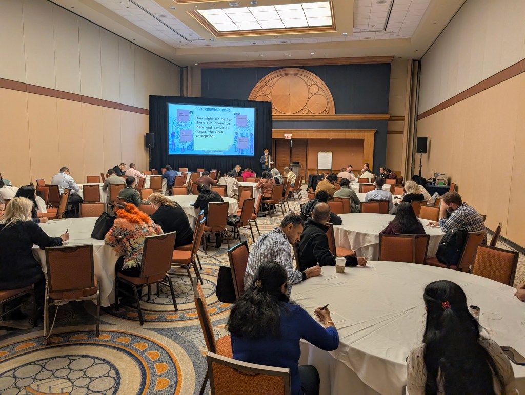 A conference room with several round tables where participants are engaged in a discussion or brainstorming session. A presentation screen is visible in the background displaying content related to crowdsourcing.