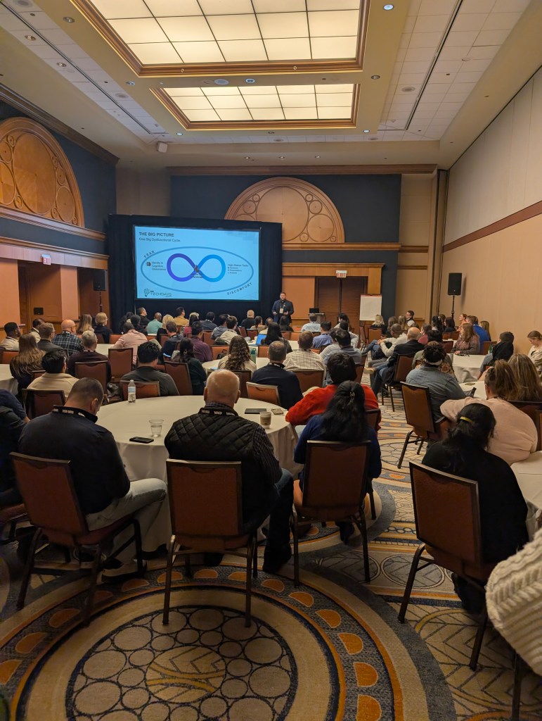 A conference presentation in a spacious meeting room with a large audience seated at round tables, focusing on a speaker in front of a projection screen displaying a diagram related to a business process.