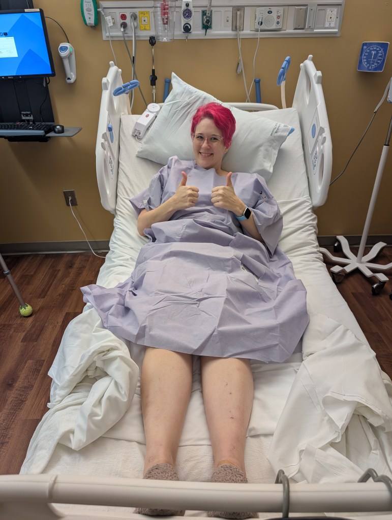 A person in a hospital gown sits on a hospital bed, smiling and giving thumbs up. The room has medical equipment and a computer in the background.