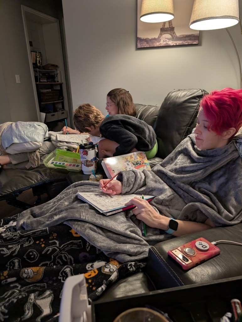 Three people sitting on a couch, wrapped in blankets, engaged in creative activities with notebooks and art supplies in a cozy living room setting.